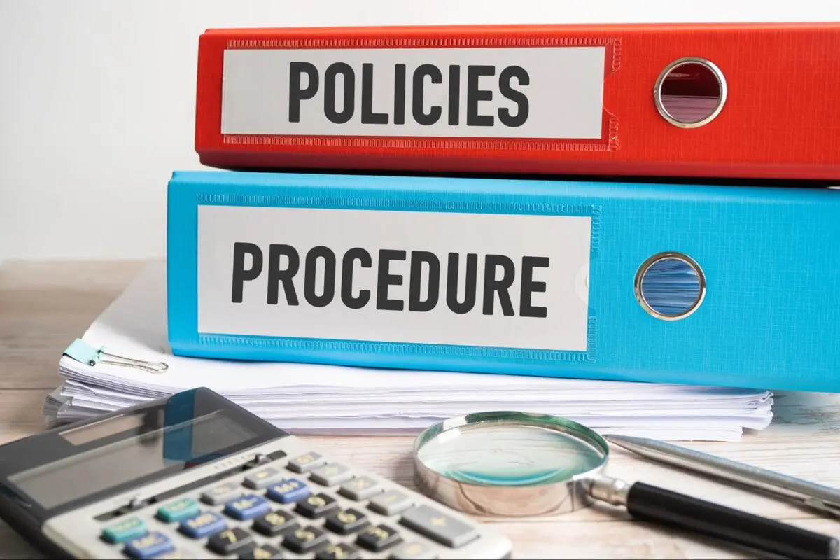 Red Policies and blue Procedure ring binders stacked on a desk with a calculator and magnifying glass