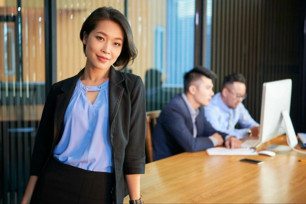 Company Secretary in a Meeting Room A businesswoman standing in a meeting room while two colleagues review documents at the table behind her