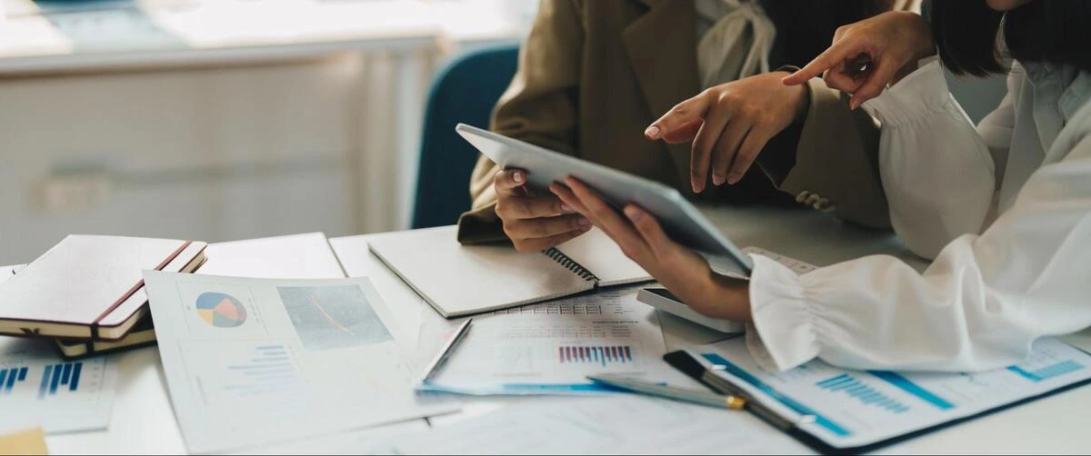 Reviewing Company Records and Reports Two people reviewing a tablet and printed financial reports spread across a desk